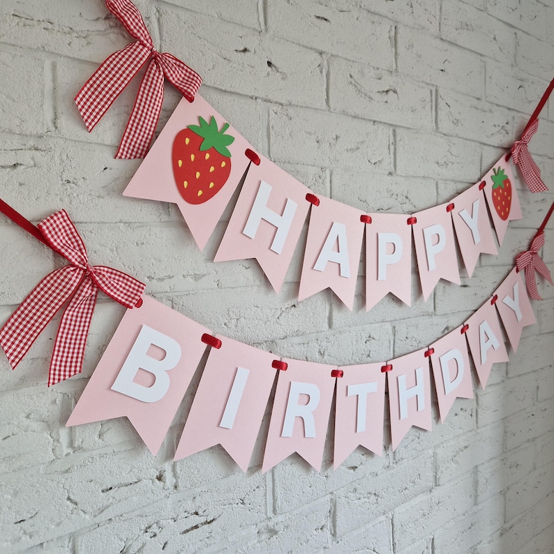 K&ouml;nnte beinhalten: Rosa Banner mit den Worten "HAPPY BIRTHDAY" in wei&szlig;en Buchstaben. Das Banner ist mit roten Erdbeeren und rot-wei&szlig; karierten Schleifen verziert. Das Banner h&auml;ngt an einer wei&szlig;en Backsteinmauer.