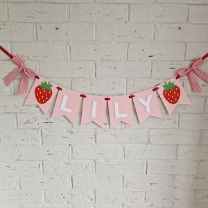 May include: Pink and white banner with the name "LILLY" spelled out in white letters. Each letter is on a separate pink triangle with a red strawberry on either end of the banner.
