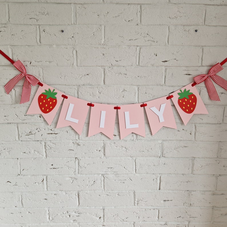 May include: Pink and white banner with the name "LILLY" spelled out in white letters. Each letter is on a separate pink triangle with a red strawberry on either end of the banner.
