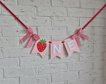 Puede incluir: Una guirnalda decorativa con tres banderines de color rosa claro colgados de una cinta roja. El primer banderín muestra una fresa roja con hojas verdes, mientras que el segundo y el tercero llevan las letras blancas N y E.