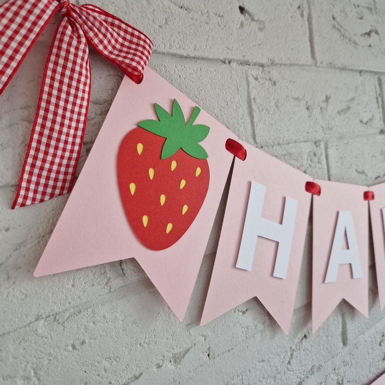 K&ouml;nnte beinhalten: Eine rosa Girlande mit einer roten Erdbeere und dem Wort "HAPPY" in wei&szlig;en Buchstaben. Die Girlande ist mit einem rot-wei&szlig; karierten Band verziert und h&auml;ngt vor einer wei&szlig;en Backsteinmauer.