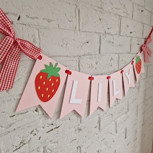 May include: Pink and white banner with the name "LILY" in white letters and a red strawberry graphic on each end. The banner is strung with red ribbon.