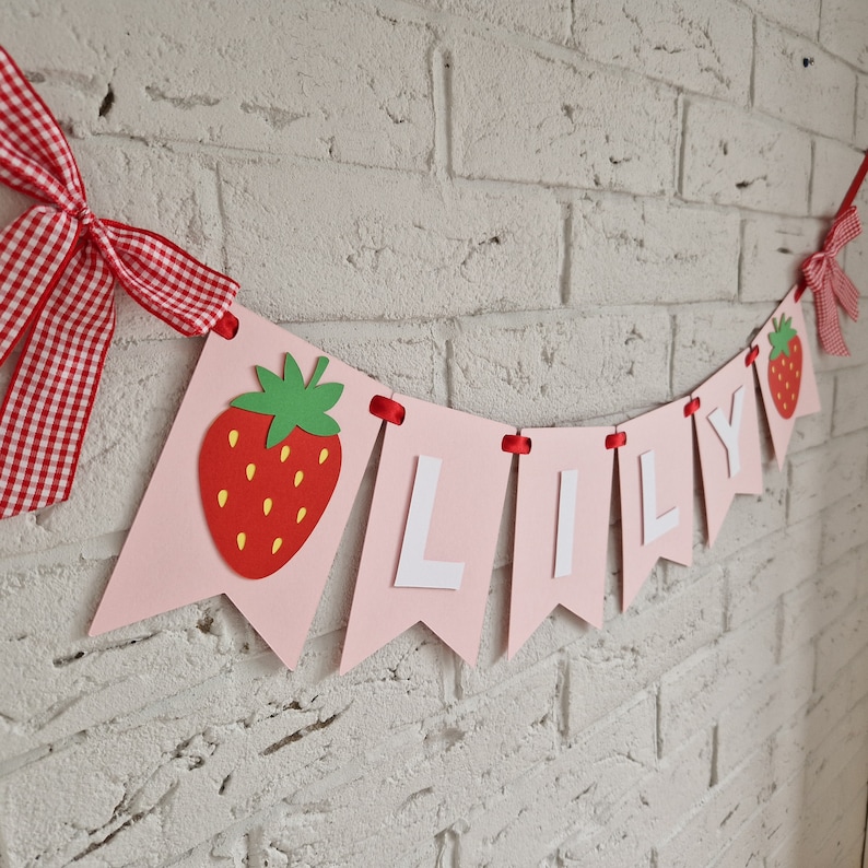 May include: Pink and white banner with the name "LILY" in white letters and a red strawberry graphic on each end. The banner is strung with red ribbon.
