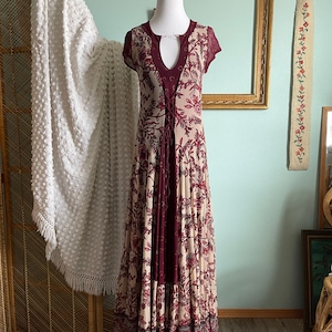 May include: A long, flowing dress with a floral pattern in cream, burgundy, and gray tones. The dress features a keyhole neckline, short sleeves, and a lace-up bodice. The dress is displayed on a mannequin.