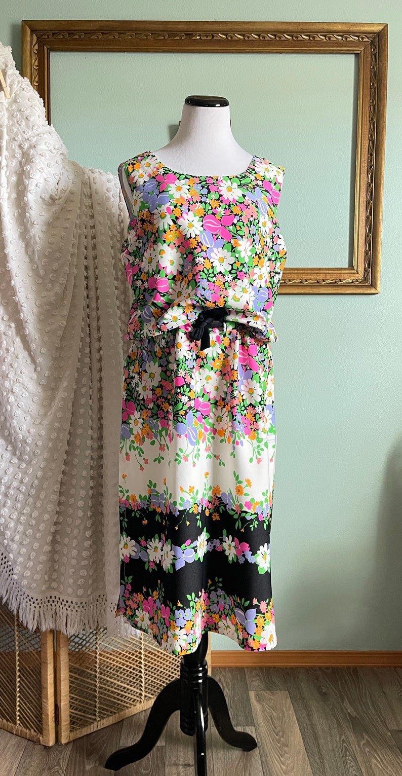 Puede incluir: Un vestido sin mangas con estampado floral en tonos rosa, morado, amarillo y verde. El vestido tiene una cinta negra atada a la cintura y un patr&oacute;n de rayas blancas y negras en la parte inferior.