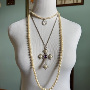 May include: A white mannequin displays three necklaces. The top necklace is a delicate strand of small pearls with a teardrop pendant. The middle necklace is a silver chain with a cross pendant featuring pearls and a purple stone. The longest necklace is a strand of cream-colored pearls.