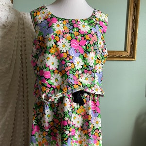 Puede incluir: Un vestido sin mangas con fondo negro y estampado floral de colores. El vestido tiene un lazo en la cintura y un dobladillo blanco.
