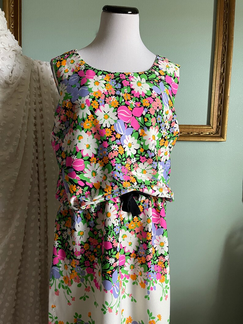 Puede incluir: Un vestido sin mangas con fondo negro y estampado floral de colores. El vestido tiene un lazo en la cintura y un dobladillo blanco.