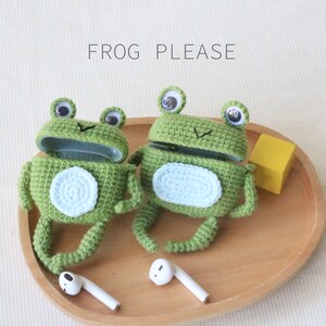 K&ouml;nnte beinhalten: Zwei gr&uuml;ne, geh&auml;kelte froschf&ouml;rmige H&uuml;llen mit Wackelaugen und hellblauen Bauchflecken. Die H&uuml;llen sind ge&ouml;ffnet und zeigen ein hellblaues Innere. Der Text "FROG PLEASE" steht &uuml;ber den Fr&ouml;schen. Zwei wei&szlig;e Ohrh&ouml;rer befinden sich vor den H&uuml;llen.