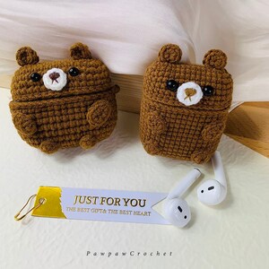 Puede incluir: Dos estuches para AirPod de color marr&oacute;n hechos a crochet con forma de oso, con ojos negros y narices blancas. Los estuches est&aacute;n sobre una superficie de color marr&oacute;n claro. Hay una etiqueta blanca con letras doradas que dice "Just for you, The best gift & the best heart." y una etiqueta blanca con el texto "Pawpaw Crochet". Tambi&eacute;n se ven dos auriculares AirPod blancos.