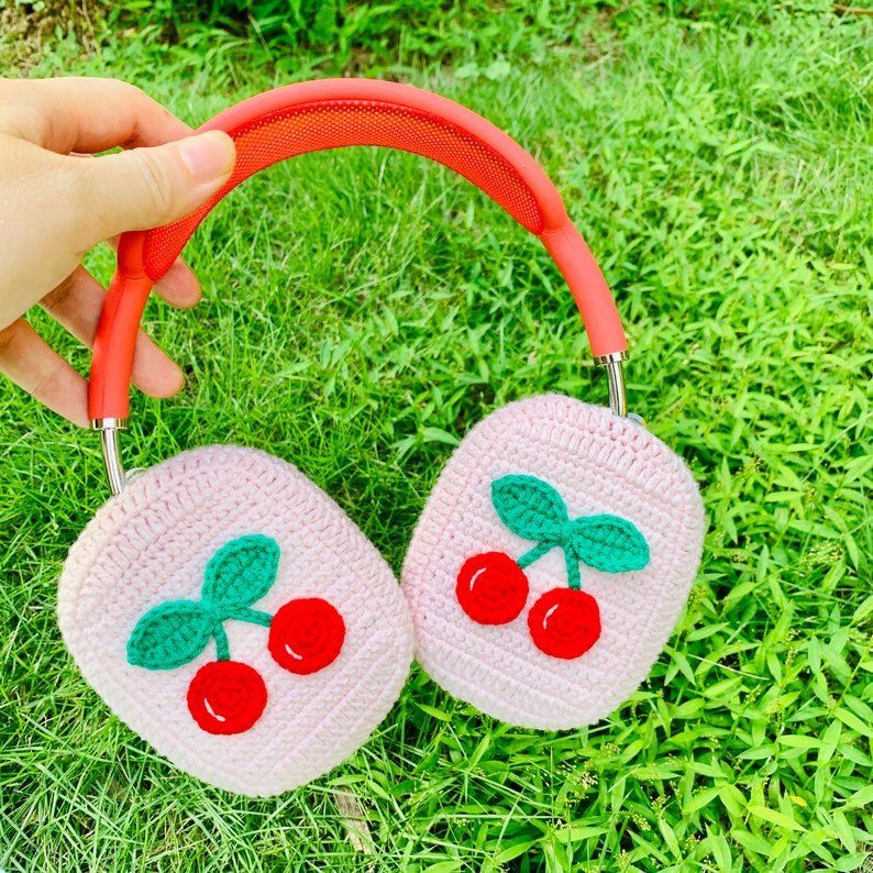 Pode incluir: Protetores de ouvido de croch&ecirc; rosa com um design de cereja. Os protetores de ouvido est&atilde;o presos a uma faixa de cabe&ccedil;a vermelha.