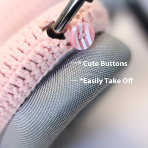 Pu&ograve; includere: Primo piano di una copertura per cuffie in crochet rosa con simpatici bottoni a righe rosa e bianchi. Il testo "*Cute Buttons" e "*Easily Take Off" &egrave; visibile nell'immagine.