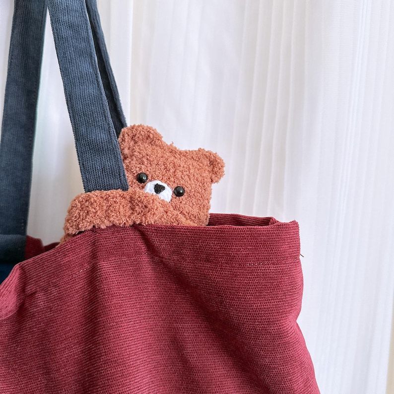 May include: A deep red tote bag with dark blue corduroy handles. A small, fuzzy brown teddy bear plush is tucked inside the bag, with its head and arms visible over the top edge. The bear has black bead eyes and a white stitched nose area.