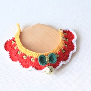 May include: Crocheted pet collar in red, yellow, and white with a green bow and gold accents. The collar has a scalloped edge and a small gold bell charm. It is designed to be worn around a pet's neck.