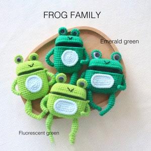 K&ouml;nnte beinhalten: Vier geh&auml;kelte Froschfiguren in Gr&uuml;nt&ouml;nen, darunter Smaragdgr&uuml;n und Leuchtgr&uuml;n. Jeder Frosch hat Kulleraugen und einen wei&szlig;en Bauch. Der Text "FROG FAMILY" ist im Bild enthalten.