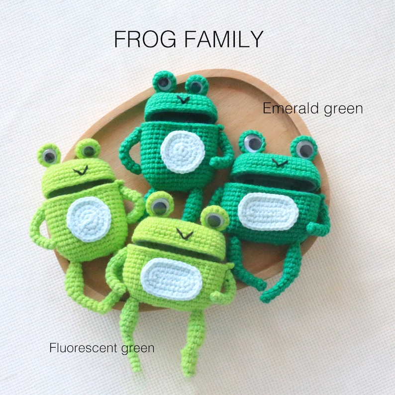 K&ouml;nnte beinhalten: Vier geh&auml;kelte Froschfiguren in Gr&uuml;nt&ouml;nen, darunter Smaragdgr&uuml;n und Leuchtgr&uuml;n. Jeder Frosch hat Kulleraugen und einen wei&szlig;en Bauch. Der Text "FROG FAMILY" ist im Bild enthalten.