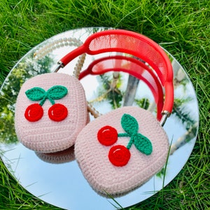 Pode incluir: Capas de auscultadores de crochet cor-de-rosa com um design de cereja. As capas s&atilde;o feitas de fio macio e apresentam uma cereza vermelha com um caule e folhas verdes.