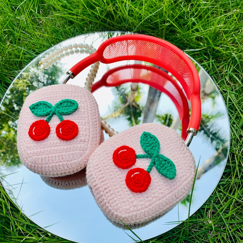 Pode incluir: Capas de auscultadores de crochet cor-de-rosa com um design de cereja. As capas s&atilde;o feitas de fio macio e apresentam uma cereza vermelha com um caule e folhas verdes.
