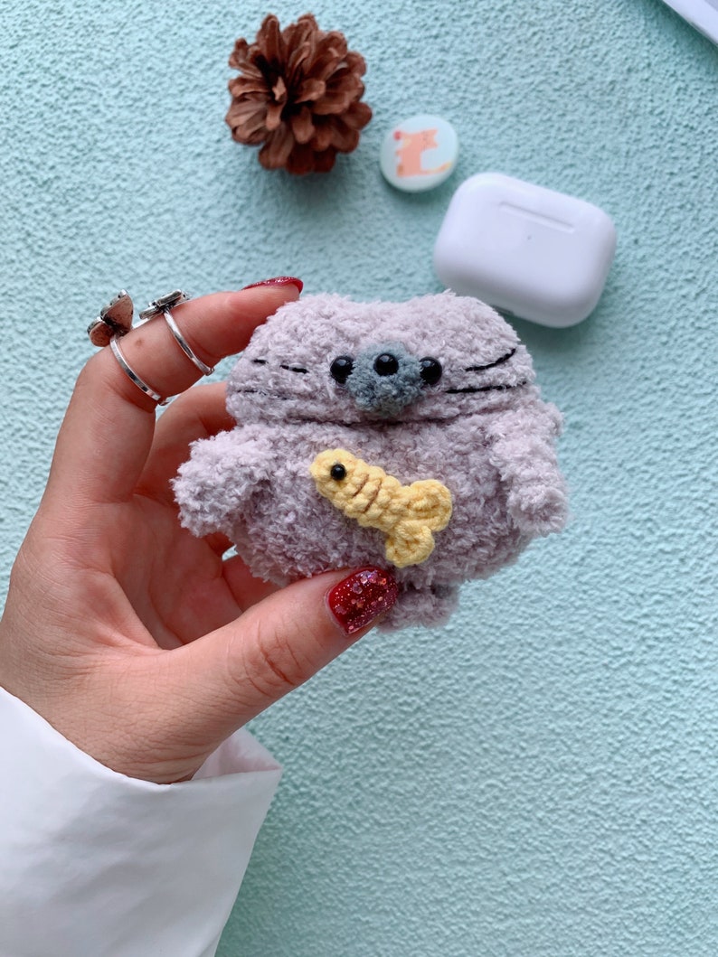 May include: A plush gray and white cat-shaped AirPods case with a yellow fish in its mouth. The case is being held in a hand with red nail polish.
