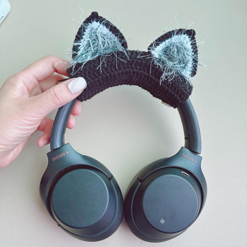 May include: Black crochet cat ear headband with fluffy gray accents, shown on a pair of black Sony headphones. The headband is held by a hand with a light-colored background.