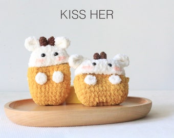 May include: Two crocheted Airpod cases shaped like cute animals with brown eyes and white fur. The cases are a light yellow color and are sitting on a wooden tray. The text "KISS HER" is written in black above the cases.
