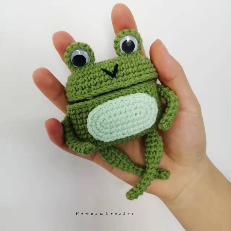 K&ouml;nnte beinhalten: Eine handgefertigte, gr&uuml;ne geh&auml;kelte Froschfigur mit gro&szlig;en Wackelaugen und einem hellgr&uuml;nen Bauch. Der Frosch ist aufwendig gestrickt. Der Text "Pawpaw Crochet" ist unten im Bild zu sehen.
