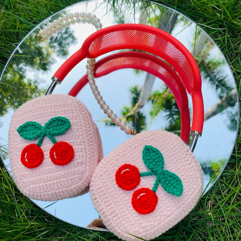 Pode incluir: Um par de capas de fone de ouvido de croch&ecirc; rosa com desenhos de cerejas. As capas s&atilde;o presas a uma faixa de cabe&ccedil;a vermelha com um colar de p&eacute;rolas.