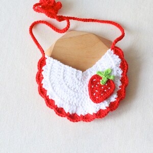 May include: A white crochet baby bib with a red scalloped edge and a red strap. The bib is decorated with a red strawberry with green leaves. The bib is designed to be tied around the neck.