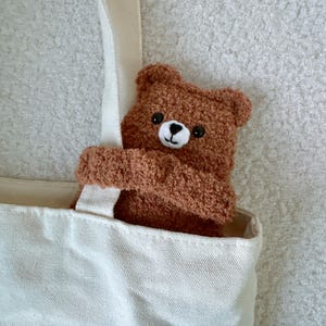 May include: A cream-colored canvas tote bag featuring a plush, brown, fuzzy teddy bear attached to the front. The bear has a white embroidered muzzle, a black stitched nose and mouth, and two small black eyes, appearing to hug the bag strap.
