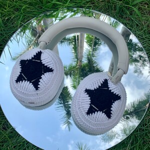May include: White headphones with black star-shaped crochet ear covers. The headphones are reflected in a mirror on a bed of green grass.