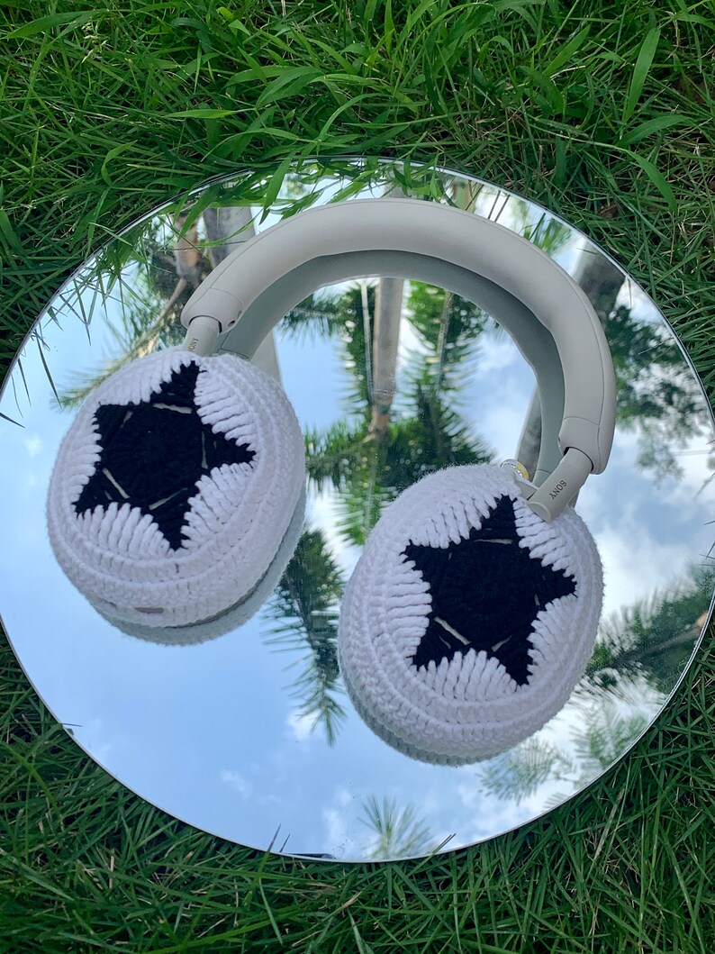 May include: White headphones with black star-shaped crochet ear covers. The headphones are reflected in a mirror on a bed of green grass.