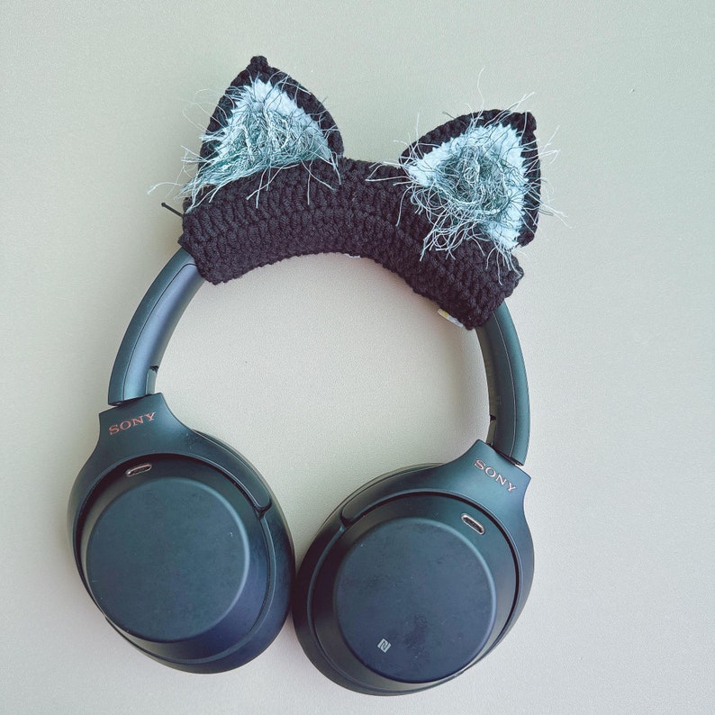 May include: Black crochet cat ear headphones accessory. The cat ears are black with silver and white fluffy accents. The headphones are dark gray with the brand name "SONY" visible on the side.