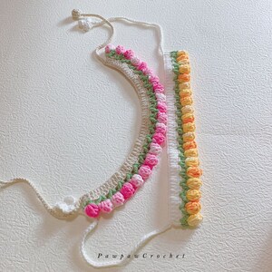 May include: Two crocheted necklaces with a flower design. One necklace is beige with pink flowers and the other is white with yellow flowers. The necklaces are on a white surface. Pawpaw Crochet.
