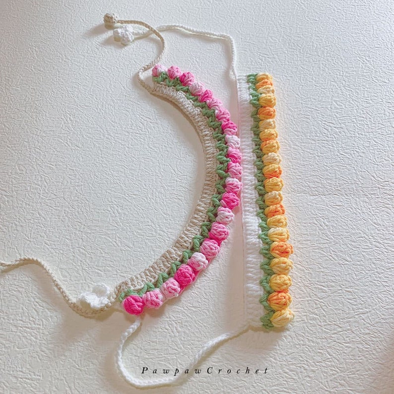 May include: Two crocheted necklaces with a flower design. One necklace is beige with pink flowers and the other is white with yellow flowers. The necklaces are on a white surface. Pawpaw Crochet.