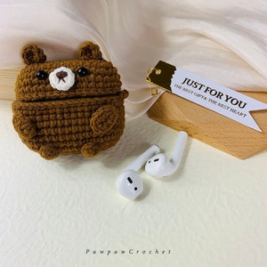 Puede incluir: Una funda de ganchillo marr&oacute;n con forma de oso para auriculares inal&aacute;mbricos. La funda est&aacute; abierta y los auriculares blancos son visibles. La etiqueta dice "Just for you, the best gift & the best heart".