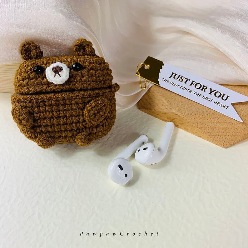Puede incluir: Una funda de ganchillo marr&oacute;n con forma de oso para auriculares inal&aacute;mbricos. La funda est&aacute; abierta y los auriculares blancos son visibles. La etiqueta dice "Just for you, the best gift & the best heart".