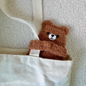 May include: A cream-colored canvas tote bag featuring a small, fuzzy brown teddy bear plush toy attached to the strap. The bear has a soft, textured surface, round ears, black eyes, and a white muzzle with a stitched black nose and mouth.