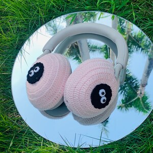 May include: Pink crocheted earmuffs with black eyes on a pair of headphones. The earmuffs are on a silver reflective surface.