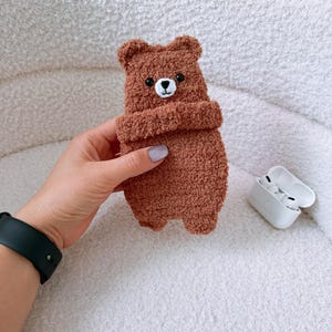 May include: A brown fuzzy bear-shaped protective case for wireless earbuds. The case features a textured, plush material, small rounded ears, and a stitched white muzzle with a black nose and two black bead eyes. The case is held in a hand.