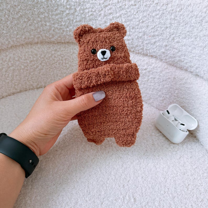 May include: A brown fuzzy bear-shaped protective case for wireless earbuds. The case features a textured, plush material, small rounded ears, and a stitched white muzzle with a black nose and two black bead eyes. The case is held in a hand.