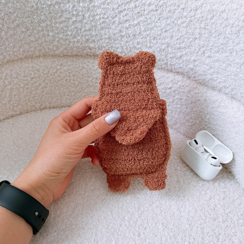 May include: A brown, fuzzy, plush-textured protective case for wireless earbuds, shaped like a bear with small rounded ears at the top and a flap-style closure on the front. The case has a soft, fleece-like material and a simple, solid brown color.