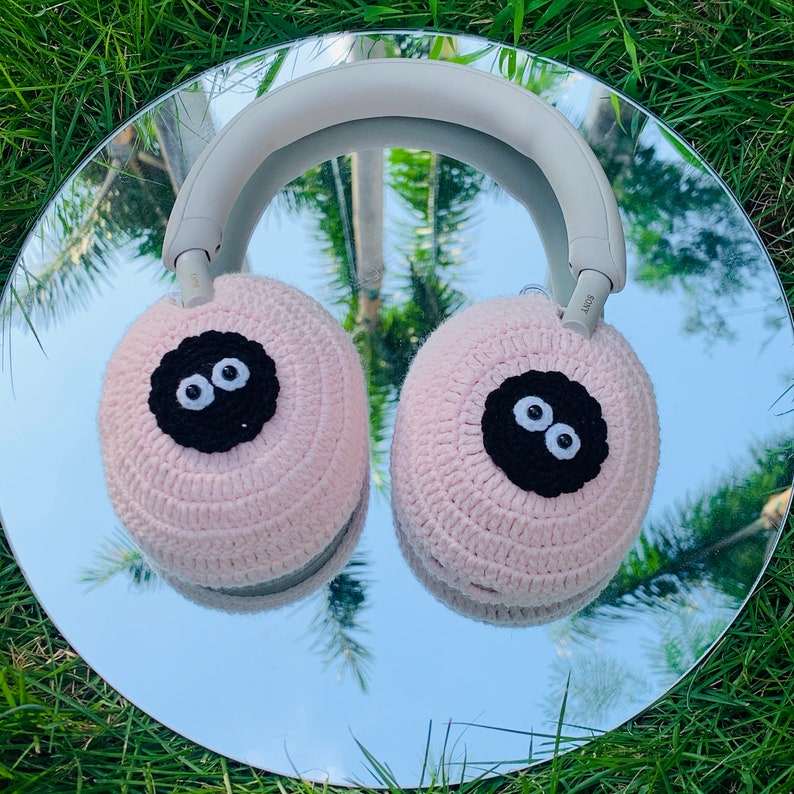 May include: Pink crocheted headphone covers with black embroidered eyes. The covers are designed to fit over the earcups of headphones.