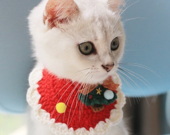 May include: A white cat wearing a red and white crocheted Christmas collar. The collar has a scalloped edge and is decorated with a small Christmas tree, a yellow circle, and a white pom-pom.