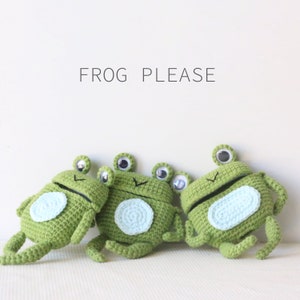 K&ouml;nnte beinhalten: Drei handgefertigte gr&uuml;ne H&auml;kelfrosch-Spielzeuge mit Wackelaugen und hellblauen B&auml;uchen. Der Text "FROG PLEASE" steht &uuml;ber den Fr&ouml;schen. Die Fr&ouml;sche sind in verschiedenen Posen, was eine verspielte Szene ergibt.