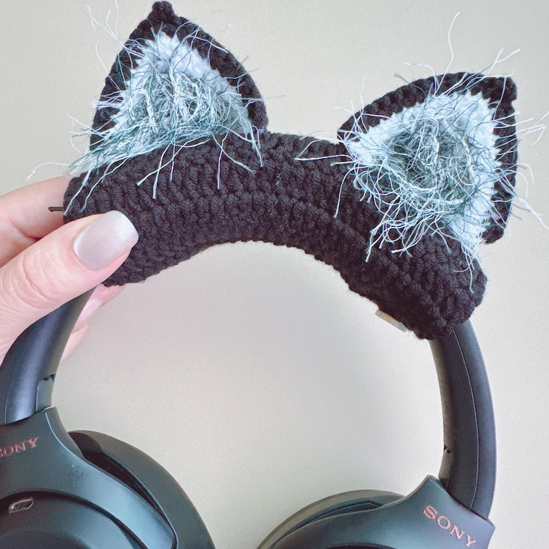 May include: Black crochet cat ear headphone covers with fluffy light blue and white accents. The cat ears are attached to a pair of black Sony headphones. The headphones are held by a person.
