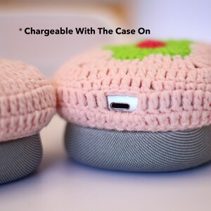 May include: Close-up of a pair of headphones with a pink crocheted cover. The cover has a green and red design on top. The headphones are gray and have a charging port. The text "Chargeable With The Case On" is visible.