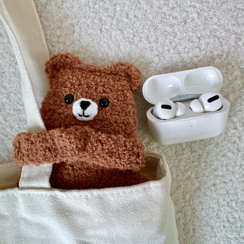 May include: A brown fuzzy bear-shaped case for wireless earbuds tucked into a cream-colored canvas tote bag, positioned next to an open white charging case containing two white wireless earbuds with black accents on the ear tips.