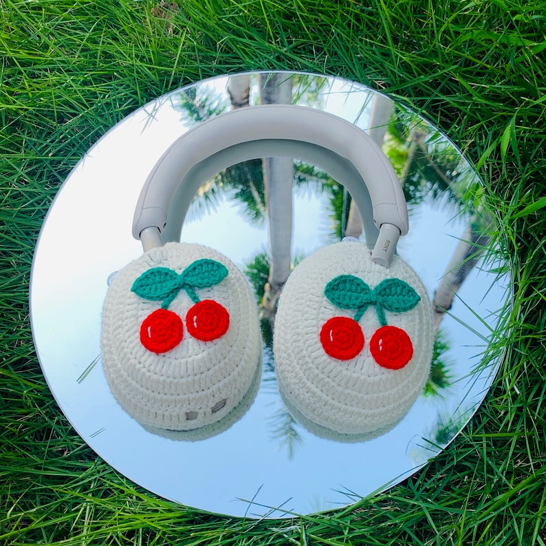 Pode incluir: Fones de ouvido de croch&ecirc; branco com um design de cereja. Os fones de ouvido t&ecirc;m uma faixa de cabe&ccedil;a branca e conchas de ouvido brancas com cerejas vermelhas e folhas verdes.