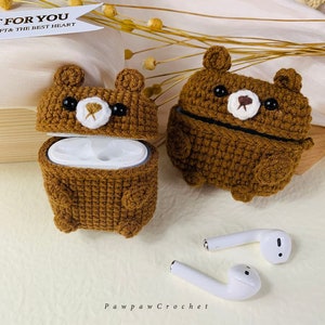 Puede incluir: Dos estuches de color marr&oacute;n hechos a crochet con forma de oso para auriculares inal&aacute;mbricos. Los estuches est&aacute;n abiertos, revelando el interior de pl&aacute;stico blanco. Dos auriculares inal&aacute;mbricos blancos est&aacute;n sobre una superficie de color marr&oacute;n claro.