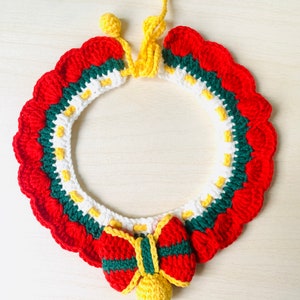 May include: A handmade crochet Christmas wreath with a scalloped edge. The wreath features red, green, white, and yellow yarn. A red bow with green and yellow stripes and a yellow ball adorn the bottom. Two yellow balls and a yellow stem are at the top.